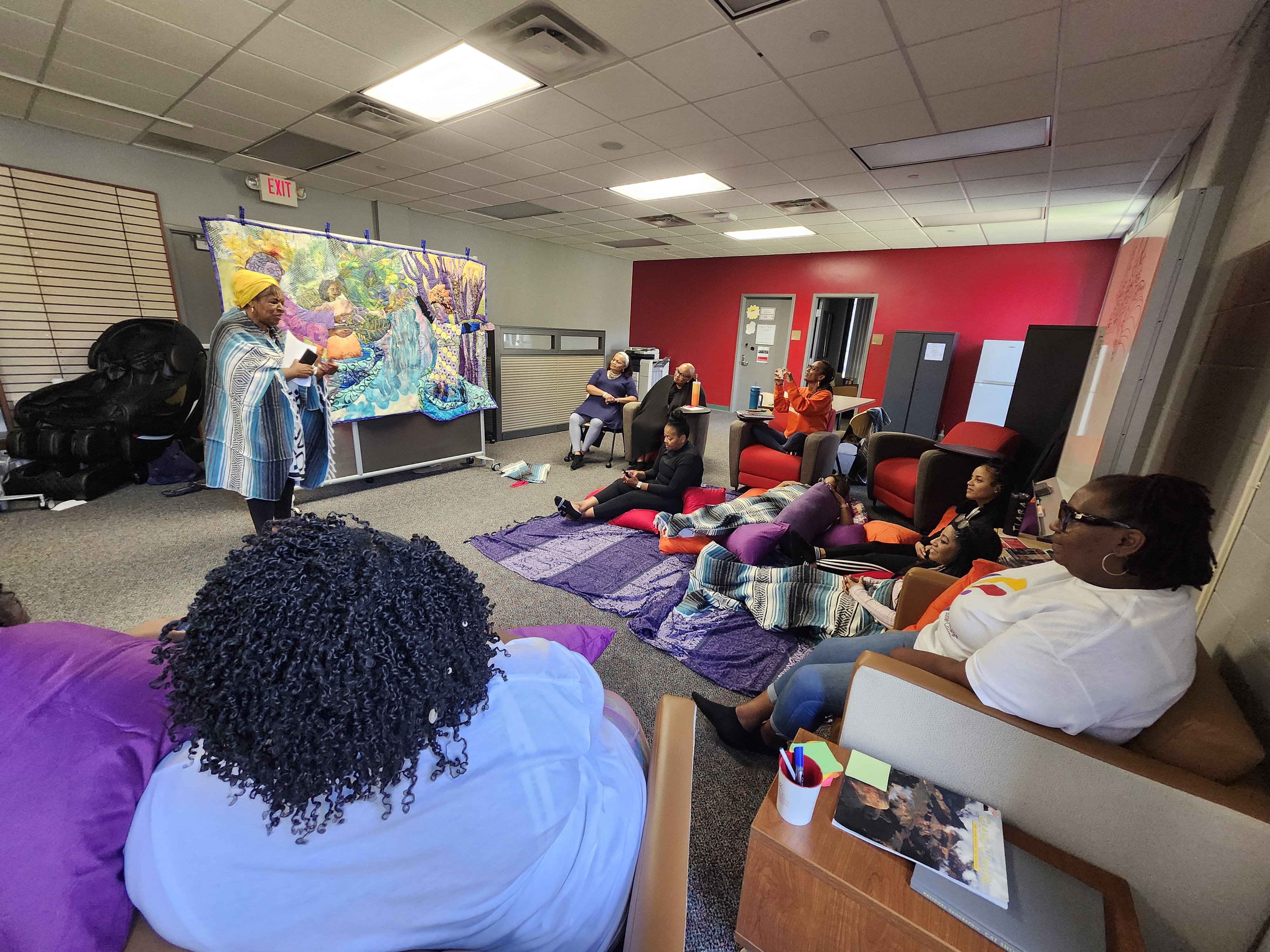 A group of people sitting in a cozy room hosting a workshop or meeting, with a woman standing and speaking in front of a colorful tapestry. Participants are engaged, some seated on the floor with blankets and cushions.
