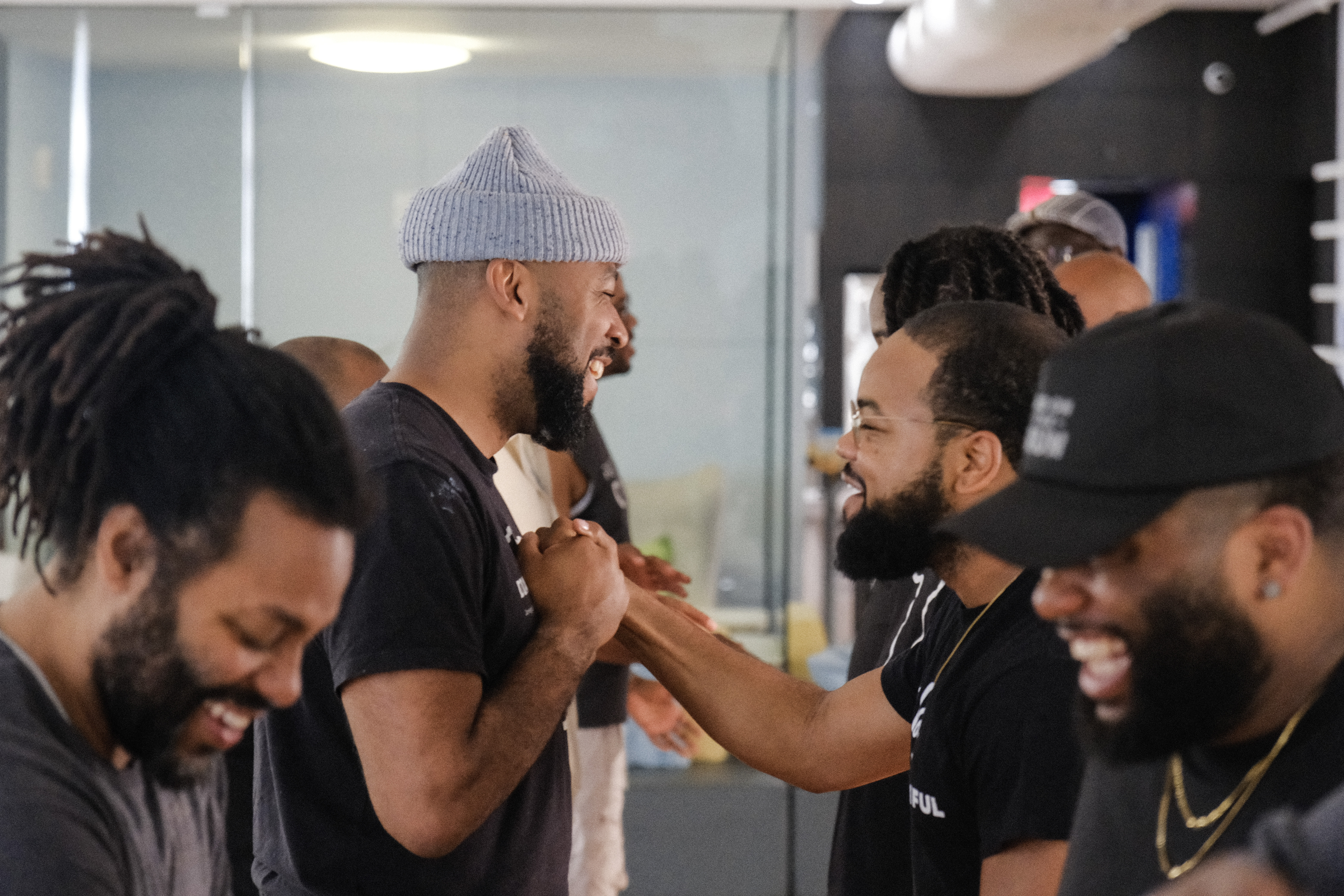 A group of men laughing and engaging in a playful interaction, shaking hands and enjoying each other's company in a bright, modern indoor setting.
