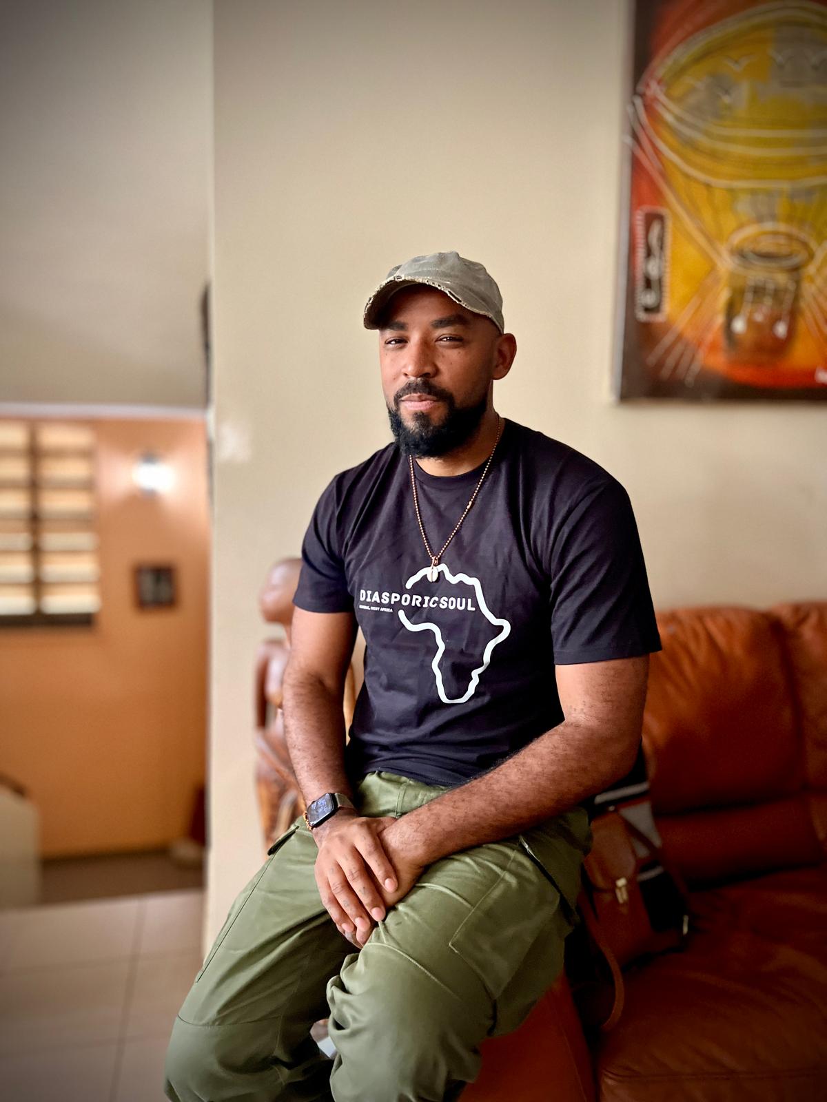 A man sitting on a sofa, wearing a black t-shirt with 'DIASPORICSoul' and Africa outline design, green cargo pants, and a light-colored cap, with a wooden sculpture and artwork in the background.