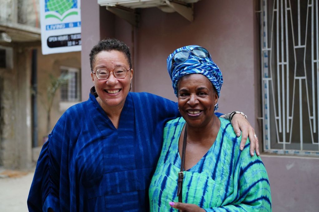 Phyllis and Angela smiling and standing side by side, wearing colorful traditional clothing after Angela hosted Phyllis in 2025 for a book talk. Phyllis who is on the left is in a blue outfit with glasses, and Angela is the woman on the right is in a blue and green outfit with a headwrap.